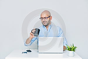 Businessman teleworking with a laptop having a coffee. Bald man with glasses