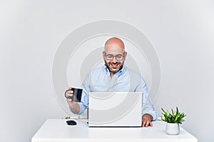 Businessman teleworking with a laptop having a coffee. Bald man with glasses