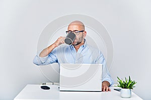 Businessman teleworking with a laptop having a coffee. Bald man with glasses