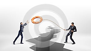 A businessman throws a lifeline with an orange life buoy over a large earthquake crack to another businessman.