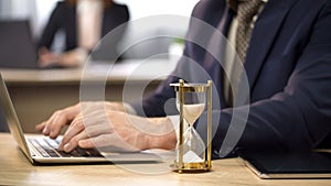 Businessman typing on laptop at desk, hourglass trickling, deadline approaching