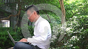 Businessman typing and working on his computer laptop in a green park, outdoors.