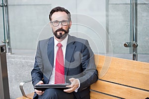 businessman using his PC tablet while sitting on bench. senior businessman using tablet computer while waiting for his car