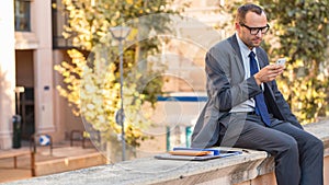 Businessman using his smart phone on a city street. He is sitti