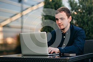 Businessman using laptop in cafe. Handsome man in dark shirn sit on the terrace. Background of green tree and blue sky