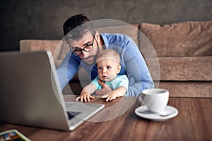 Businessman using a laptop computer for work at home while looking after his baby son.