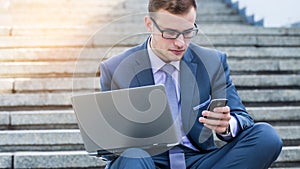 Businessman using laptop pc and mobile phone. He is sitting on a stairs.