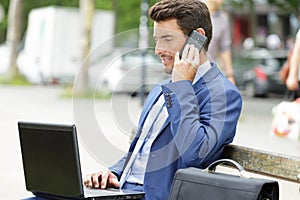 businessman using laptop and smartphone on bench outdoors