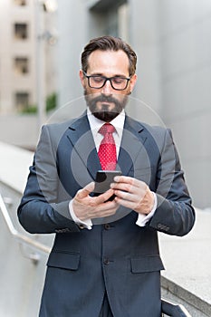 Businessman using mobile phone app reading outside of office in city with skyscrapers buildings in background.