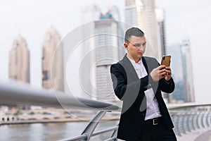 Businessman using mobile phone app texting outside of office in urban city with skyscrapers buildings in the background. Young