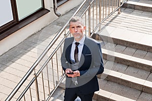 Businessman using mobile phone app texting outside of office in urban city with skyscrapers buildings in the background. Young