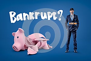 A businessman with a very tight belt, hands in pockets, standing next to a big broken piggy bank with the title