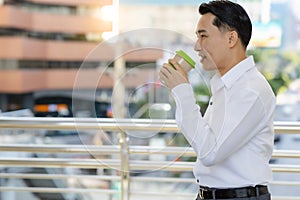 Businessman walking at city street and drinking coffee to go outside, man wearing white shirt and enjoying his hot cappuccino