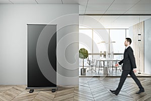 Businessman walking in modern coworking office interior with empty black banner, city view, equipment and furniture, law and legal