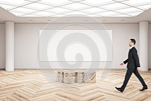 Businessman walking in modern exhibition hall interior with wooden flooring, shiny golden seat and empty mock up poster on