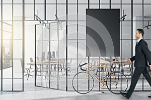 Businessman walking in modern glass partition and bike in stylish coworking office interior with empty black mock up poster and