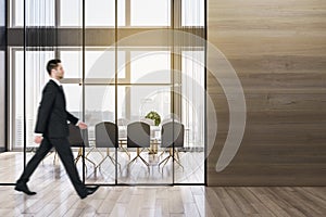 Businessman walking in modern wooden meeting room office interior with empty mock up place on wall, table, armchairs, window with