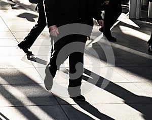 Businessman walking quickly on city sidewalk wearing black suit silhouetted in the sun with long dark shadows and bright light