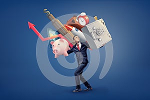 A businessman walks under a heavy weight of many things on his shoulders: a safe box, a piggy bank, skyscraper and an