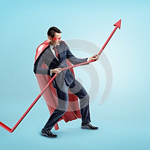 A businessman wearing a red superhero cape trying to hold a red statistic arrow with force on blue background.