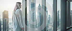 Businessman wearing traditional white outfit standing in modern office looking out on big city with skyscrapers