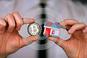 A businessman in a white shirt and black tie holds a bitcoin and miniature house in his hands