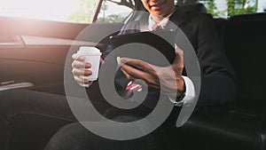 Businessman working in a car and using a tablet while drinking coffee