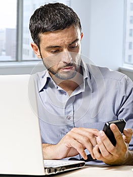 Businessman working on computer laptop using mobile phone at office desk in front of skyscraper window