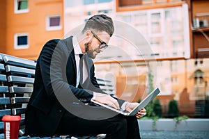 Businessman working with his computer and drinking coffee on a bench outdoors