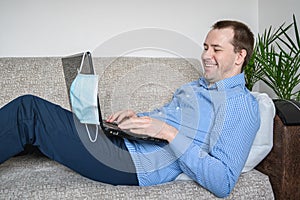 Businessman working from home, smiling. Caucasian adult man in a blue shirt sitting on a sofa, working using a laptop