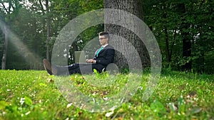 Businessman working on laptop, sitting on grass in park, escaping office routine