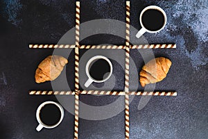 Businessman working morning with cups of hot coffee and sweet croissants on black background. Game of noughts and crosses.