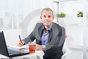 Businessman working in office, sitting at table with a laptop, looking smiling.