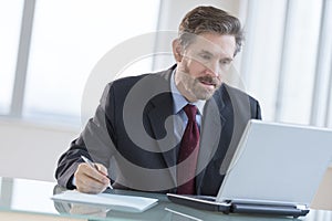 Businessman Writing Notes While Using Laptop At Desk