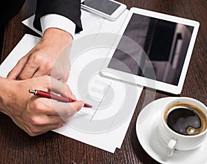 Businessman writing on the paper next to the tablet, coffee, cell phone