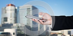 Businessmans Hand Holding Smartphone With Business City and Corporate Buildings In Background