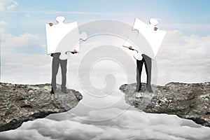 Businessmen holding jigsaw puzzles to connect on cliff with cloudscape