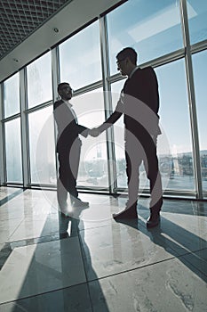 Businessmen are shaking hands with each other standing against panoramic windows.