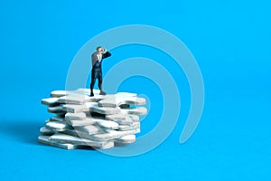 Businessmen standing above jigsaw puzzle piece stack using binoculars above wooden table. Miniature tiny people toys photography.