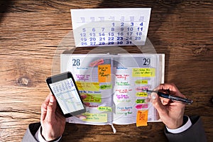 Businessperson With Mobile Phone Writing Schedule In Diary