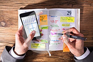 Businessperson With Mobile Phone Writing Schedule In Diary