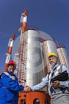 Businessperson and Worker in Unfiofm Shaking Hands Against Power Plant Chimneys and Tanks