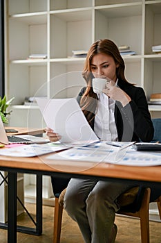 A businesswoman analyzing business financial reports while sipping coffee at her desk