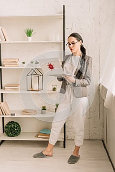 Businesswoman in blazer over pajamas using laptop with coronavirus bacteria sticker while standing near rack with books