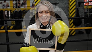 A businesswoman in a boxing gloves holds a board with the inscription WINNER.