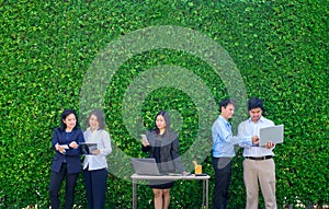 Businesswoman and businessman working outside office using laptop computer mobile phone and tablet device at green leaf