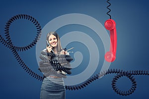 A businesswoman caught and bound by large black telephone cords near a giant red phone receiver.