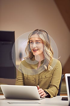 Businesswoman In Cubicle Wearing Headset Talking To Caller In Busy Customer Services Centre