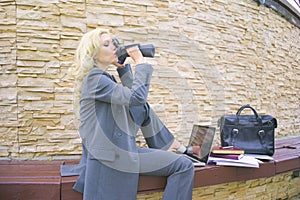 Businesswoman drinking water from a bottle. A woman secretly works at a laptop outside on a bench