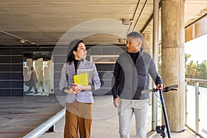 Businesswoman in formal suit talking to businessman holding a scooter. Girl in formal suit talking to man holding electric scooter